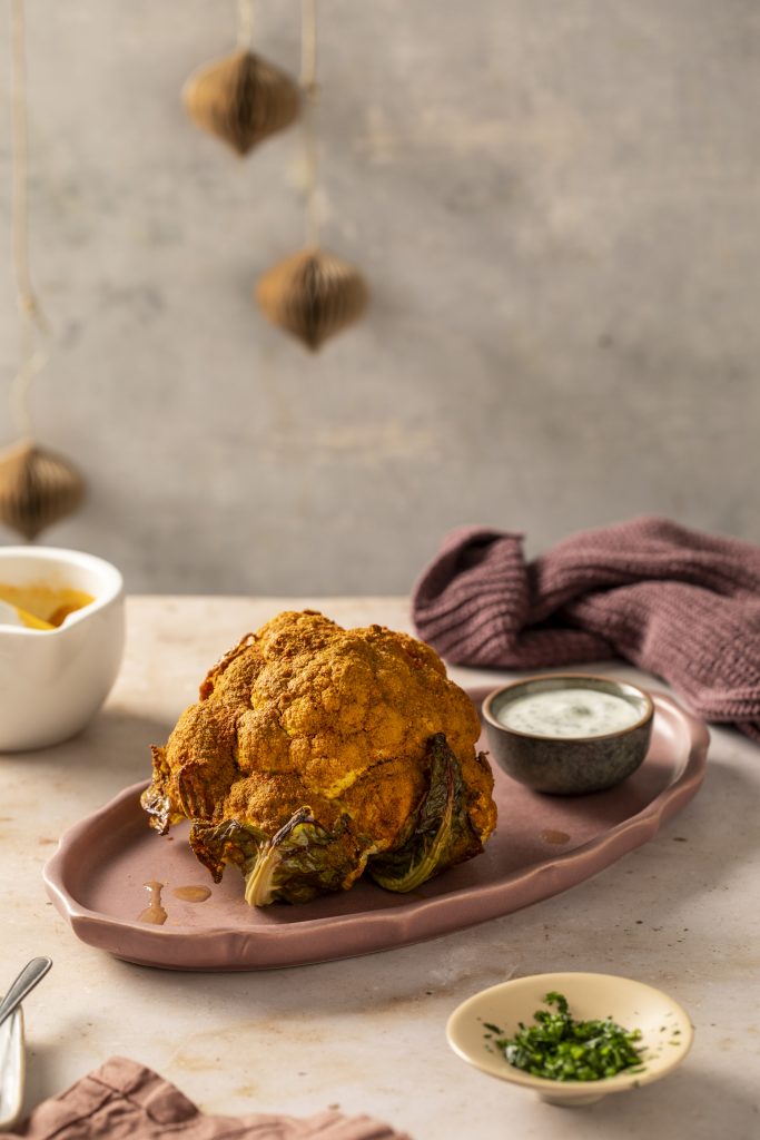 Racions: Per a 2 persones Ingredients: • 1 coliflor blanca sencera • 250 g de iogurt natural sense sucre • 1 cullerada de curri • 1 cullerada de pebre vermell fumat • Julivert fresc (al gust) • Llavors de comí (opcional) • Sal (al gust) • Oli d’oliva verge extra (quantitat necessària) • 1 cullerada d’aigua Preparació: 1. Preparar el marinat: en un bol, barrejar el curri, el pebre vermell fumat, les llavors de comí (si s’utilitzen), una mica de sal, un raig d’oli d’oliva i una cullerada d’aigua. Remenar bé fins a obtenir una pasta d’espècies. 2. Marinar la coliflor: untar tota la superfície de la coliflor amb aquesta barreja, escampant bé amb les mans perquè el condiment entri per tots els racons. 3. Enfornar: posar la coliflor en una safata i enfornar-la en forn estàtic preescalfat a 180 °C durant 35-40 minuts. Comprovar de tant en tant per evitar que es cremi. L’interior ha de quedar tendre i l’exterior daurat. 4. Preparar la salsa: mentre la coliflor es refreda una mica, barrejar en un bol el iogurt amb un raig d’oli d’oliva, una mica de sal i julivert fresc ben picat. 5. Servir: tallar la coliflor en rodanxes gruixudes i servir-la amb la salsa de iogurt i julivert per sobre o al costat.